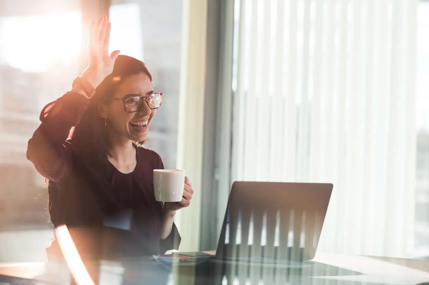 Leende person på jobbet med laptop och kaffekopp räcker upp en hand i hälsning till en kollega.