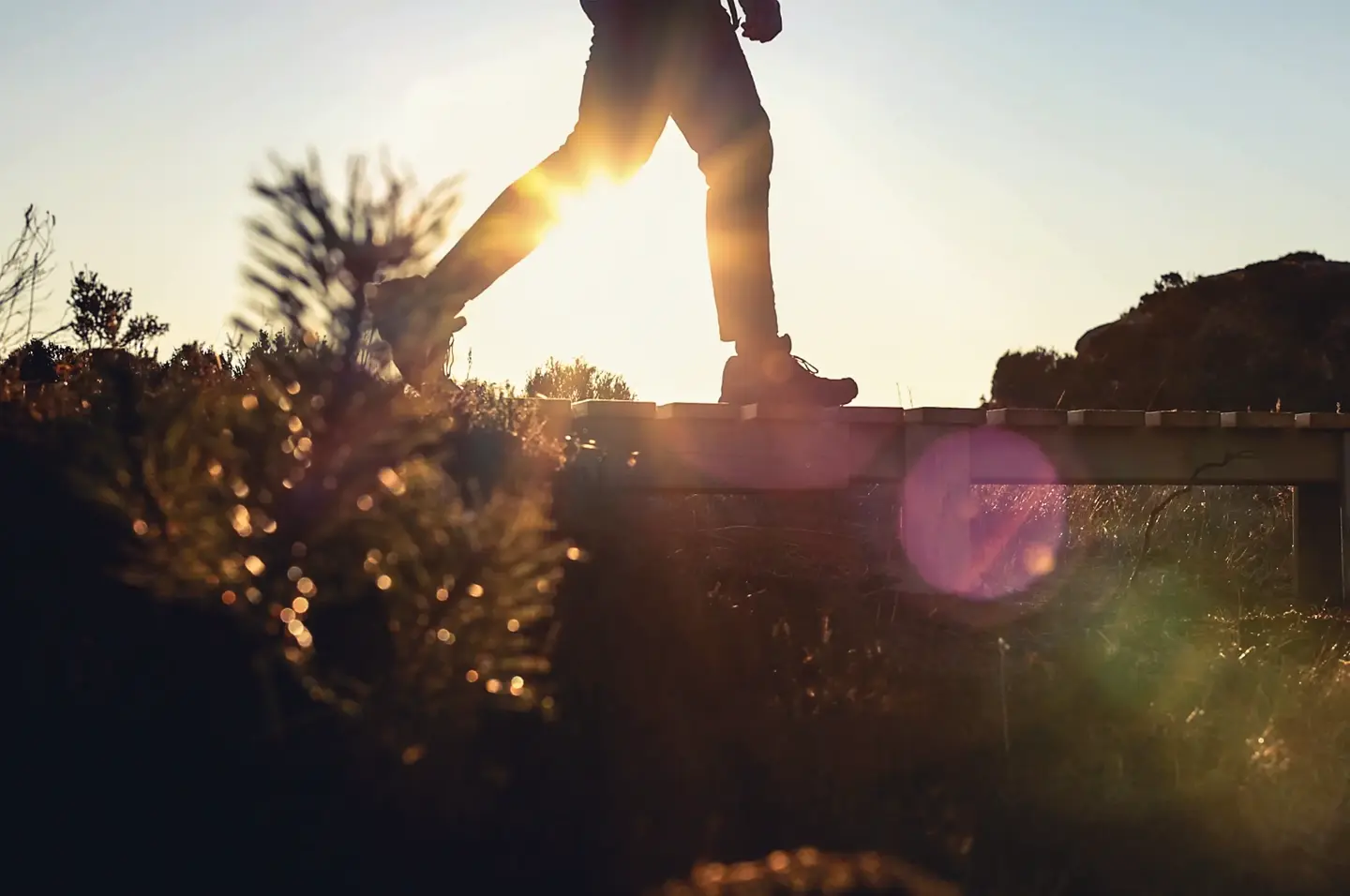 En person i vardagskläder går en promenad i det öppna landskapet och korsar en bäck över en enkel träbro.