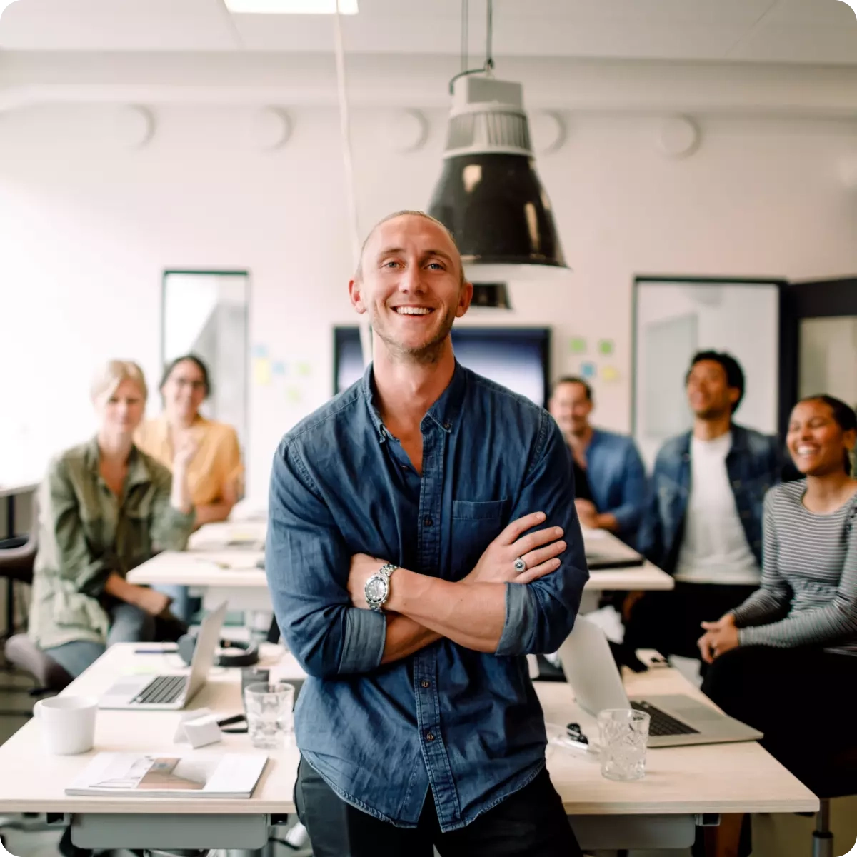 En nöjd chef ler mot kameran i kontorsmiljö med hela sitt team bakom sig.