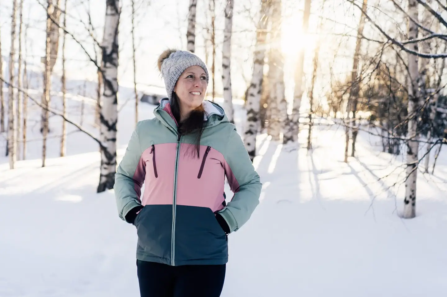 Leende person på en promenad i björkskog med snö och låg vintersol.